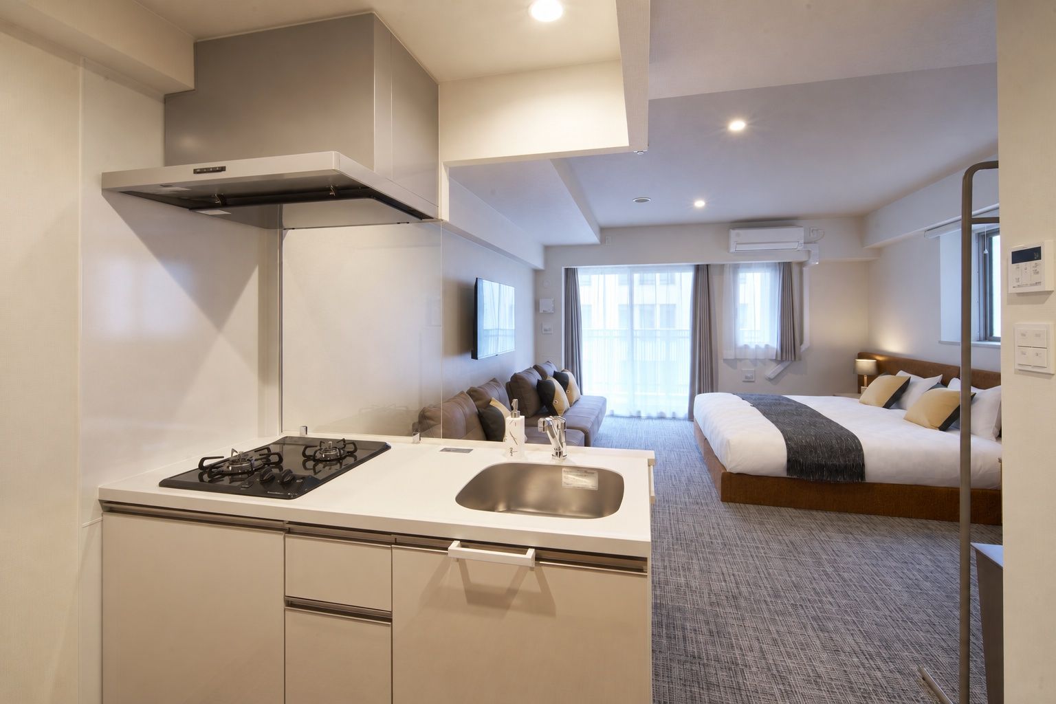 A modern hotel room with a kitchenette, a bed, a TV, and a window with curtains.