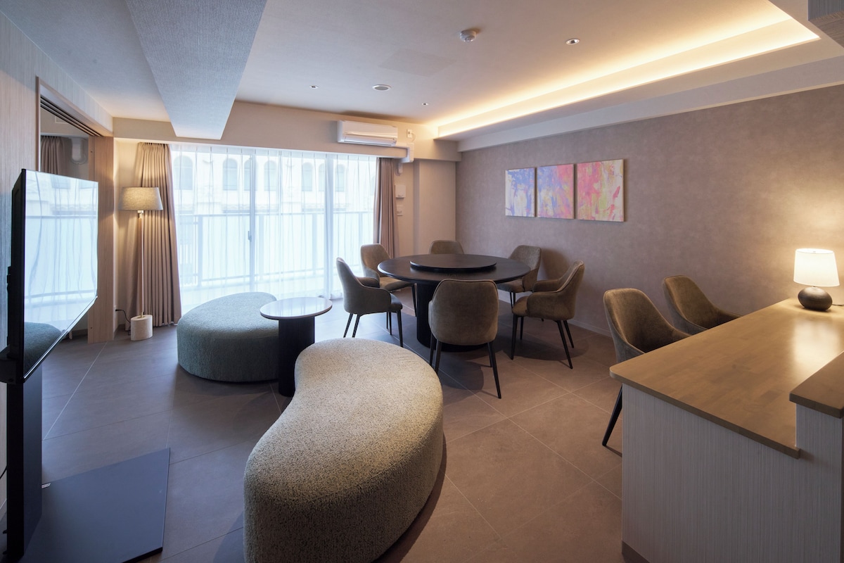 A modern hotel room with a flat-screen TV, a round dining table with chairs, a sofa, and a small kitchenette area.