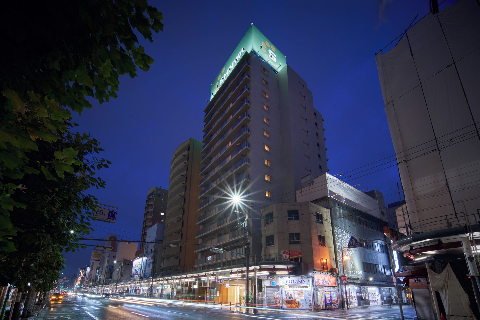 A multi-story hotel with a prominent green-lit corner, a ground-level commercial area with various shops, and a busy street with light trails indicating traffic.