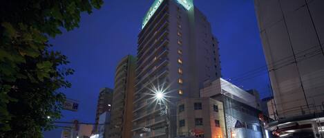 A multi-story hotel with a prominent green-lit corner, a ground-level commercial area with various shops, and a busy street with light trails indicating traffic.