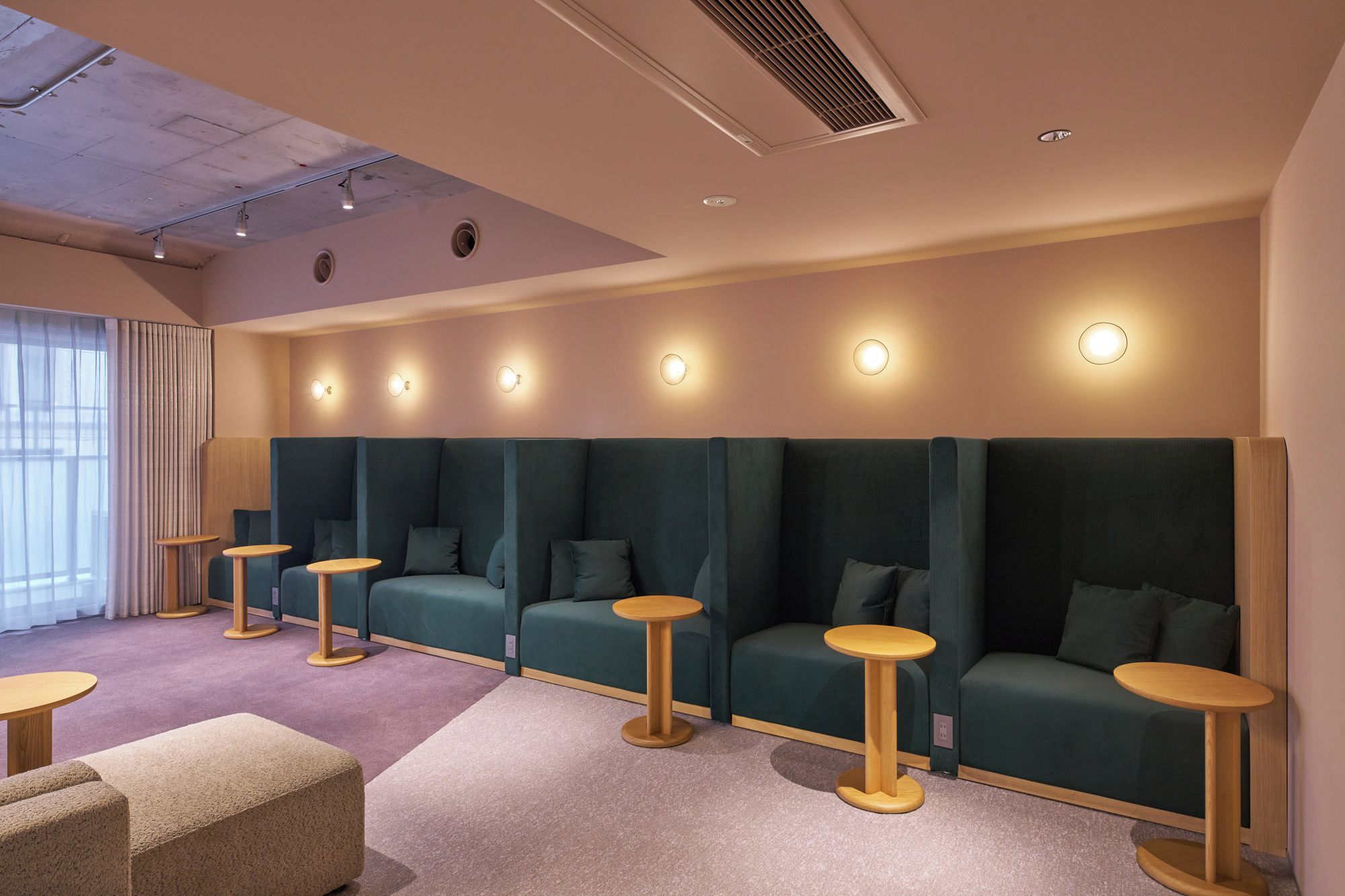 A row of green cushioned seating with round wooden tables, a ceiling with recessed lighting, and a window with sheer curtains.