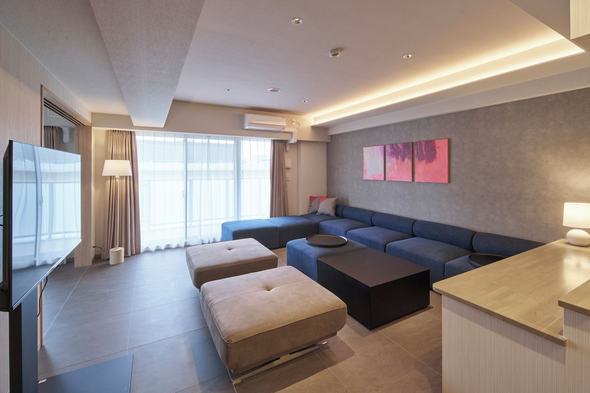 A modern hotel room with a large sofa, a flat-screen TV, a coffee table, and a desk area.