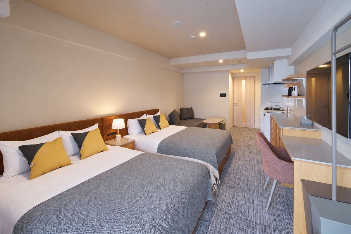 A hotel room with two beds, a desk, a chair, a small table, and a television.