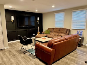 A modern living room with a brown leather sofa, a flat-screen TV, a guitar, and a small table with a plant. - Luxury Lincolnwood 3BR 2BA furnished midterm. Utilities and wifi included. (Lincolnwood)