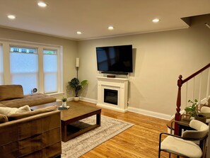 A living room with a fireplace, a television mounted above it, a sofa, a coffee table, and a staircase with a railing. - Luxury Lincolnwood 3BR 2BA furnished midterm. Utilities and wifi included. (Lincolnwood)