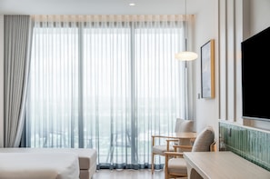 Quarto de hotel com cama, escrivaninha, duas cadeiras, televisão e cortinas translúcidas.
