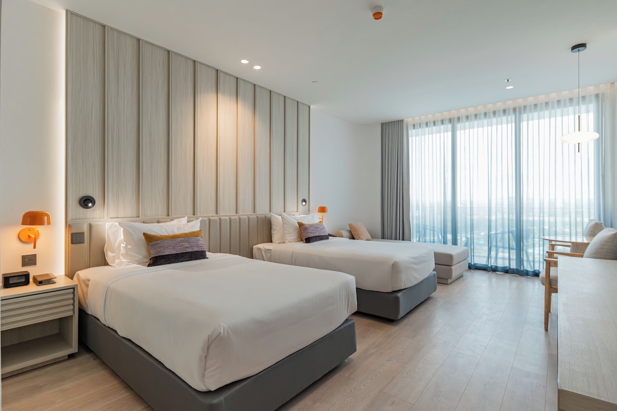 Quarto de hotel com duas camas, uma escrivaninha e uma janela grande com cortinas.