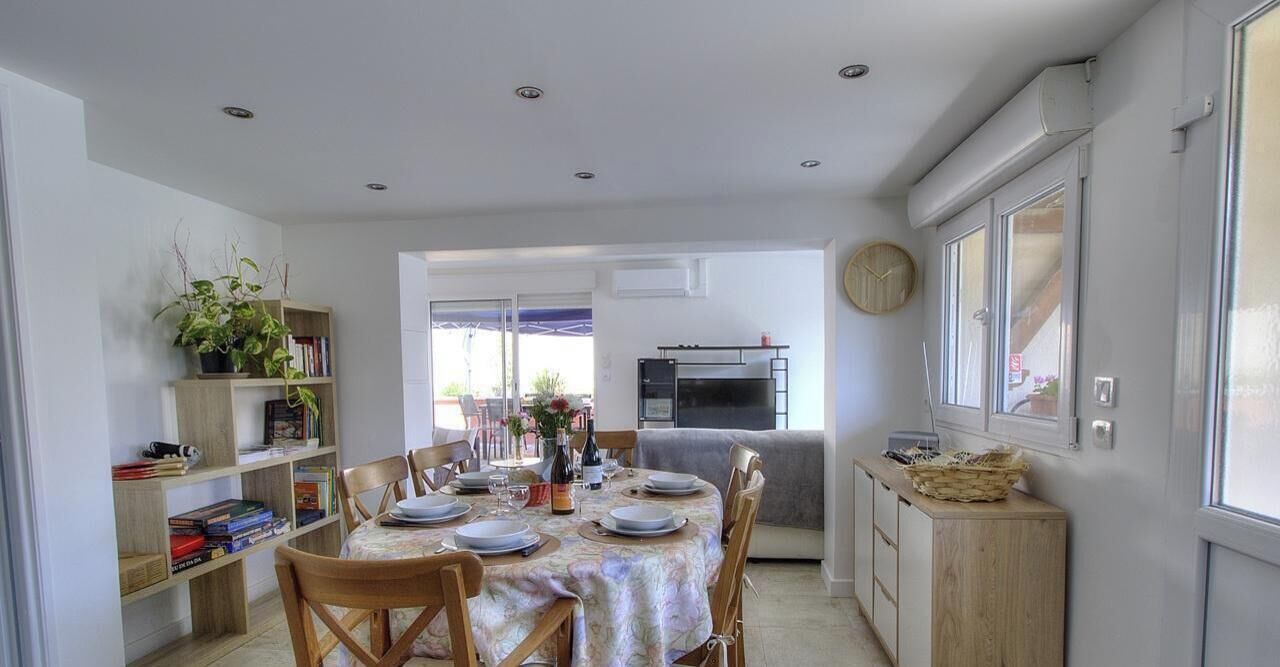Comedor con mesa lista para cuatro, rodeada de sillas, una estantería y una zona de cocina con refrigerador y horno.