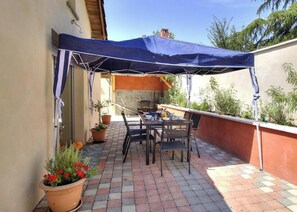 Een terras buiten met een tafel en stoelen, een blauw zonnescherm, potplanten en een tegelvloer.