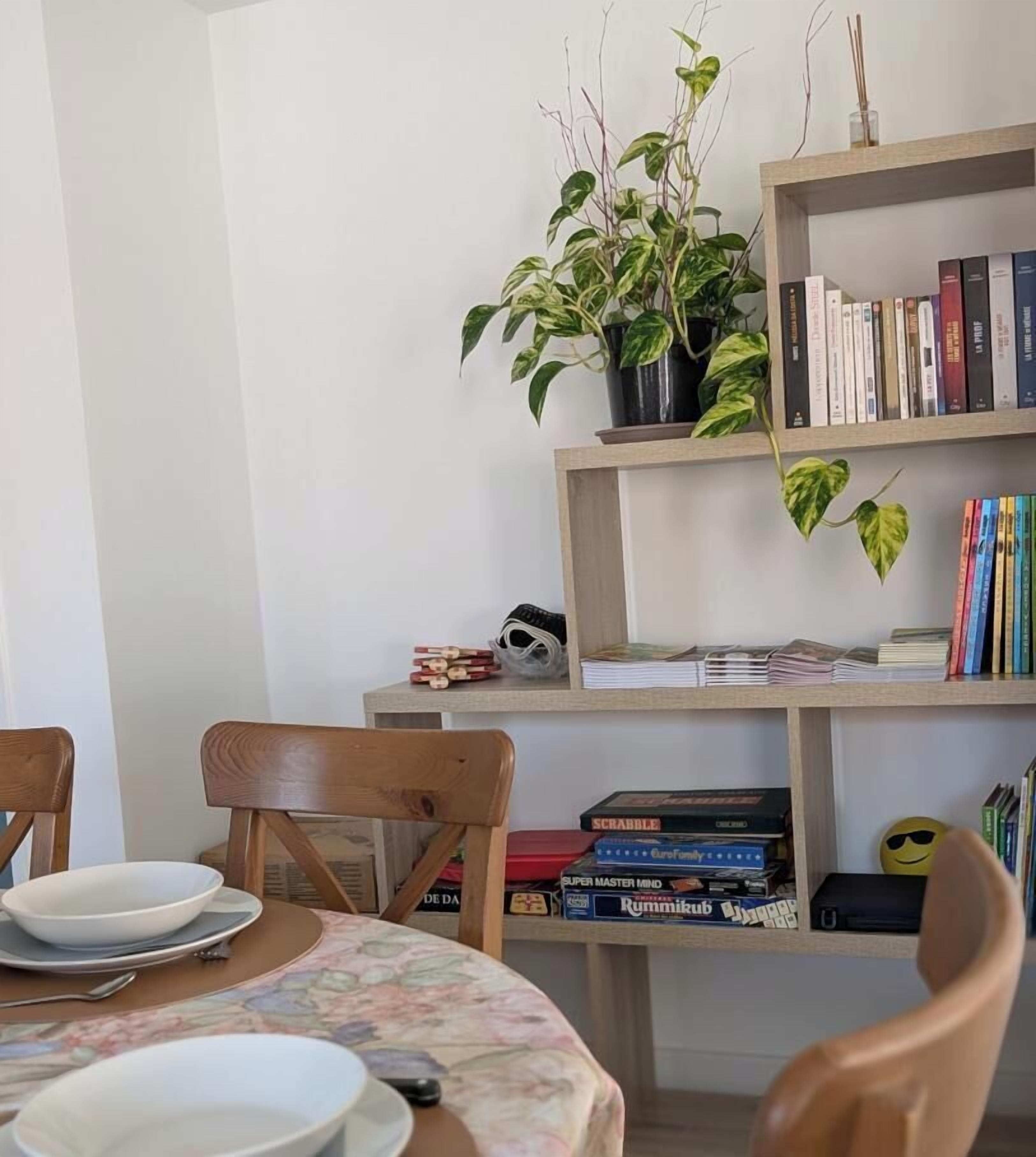 Een eethoek met een tafel gedekt voor twee personen, stoelen, een boekenkast met boeken en spellen, en een potplant.
