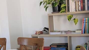 Een eethoek met een tafel gedekt voor twee personen, stoelen, een boekenkast met boeken en spellen, en een potplant.