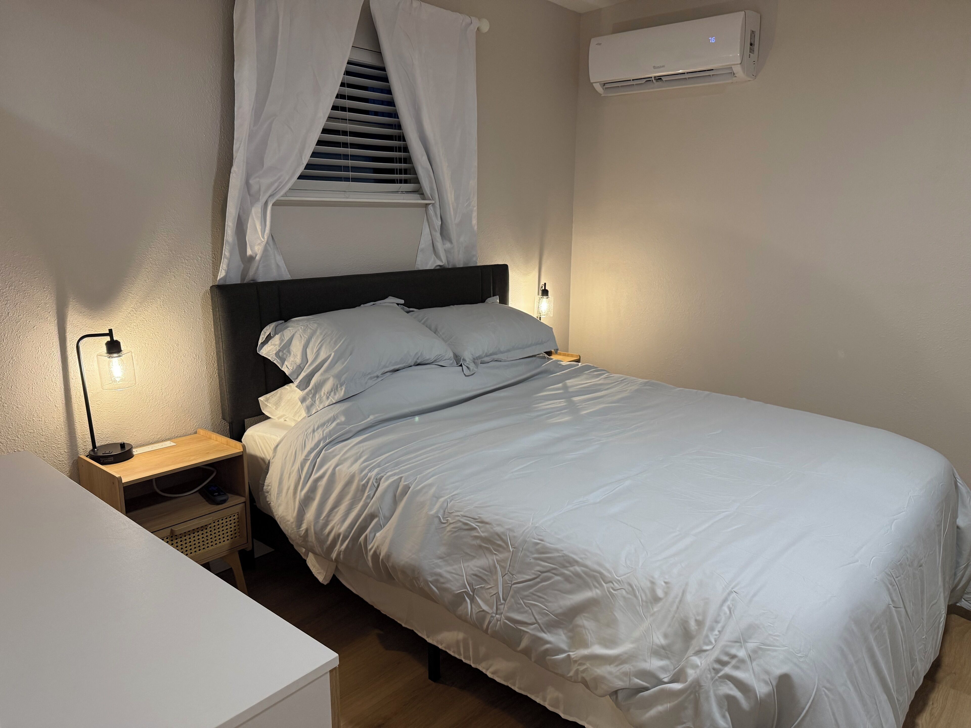 A neatly made bed with white bedding, a bedside lamp, a nightstand, and a wall-mounted air conditioner.