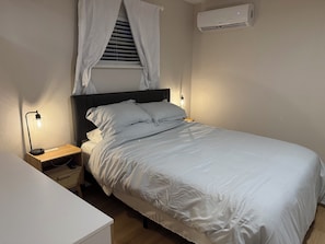 Un lit bien fait, avec une literie blanche, une lampe de chevet, une table de chevet et un climatiseur fixé au mur.