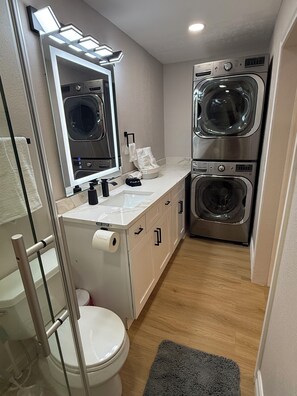 A compact bathroom with a toilet, sink, mirror, and stacked washing machines. - Modern 1BR Tampa Stay Near Downtown (Tampa)