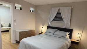 Quarto com cama, mesas de cabeceira, cômoda, espelho e janela com cortinas.