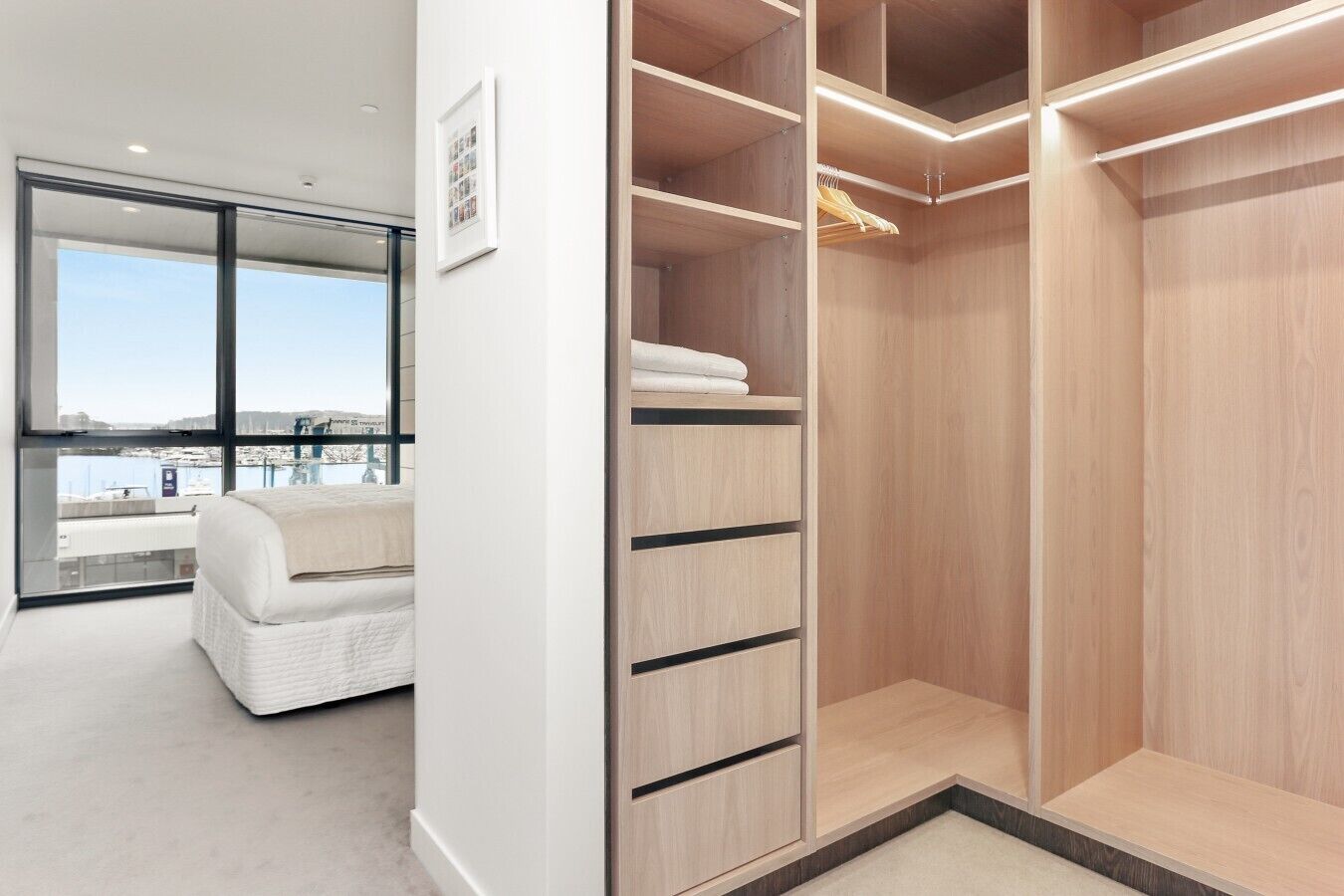 A modern bedroom with a large window, a white bed, and a walk-in closet with wooden shelving.