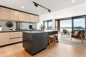 A modern kitchen with a large island, bar stools, and a dining area with a view. - Inner-City Comfort at Beaumont in Wynyard Quarter (Auckland)