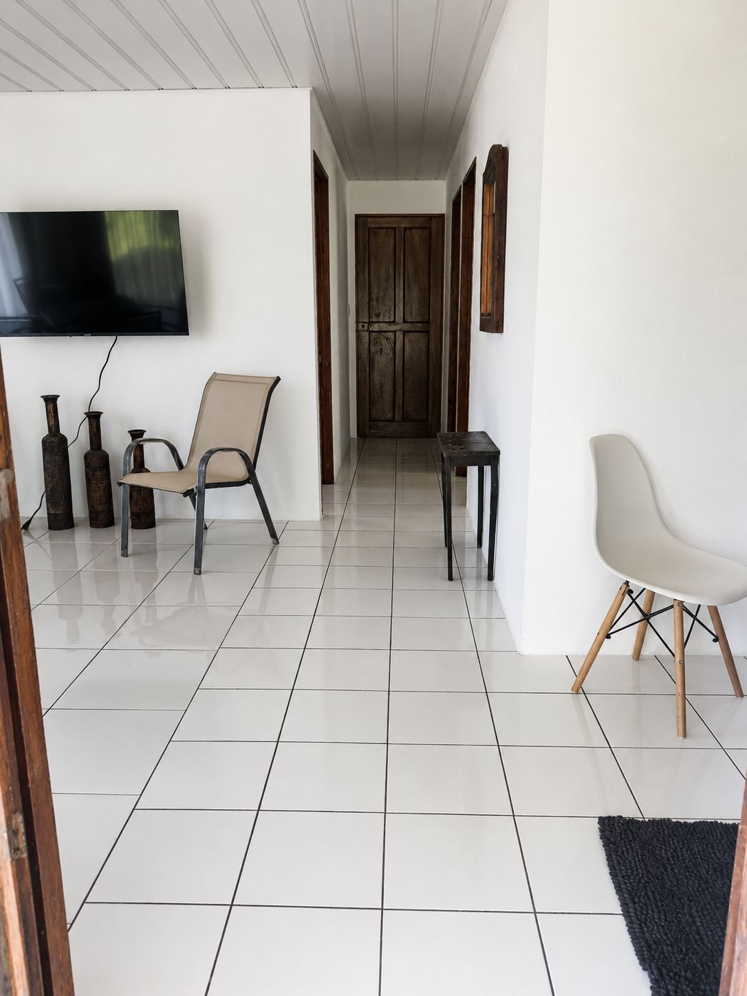 Un pasillo con un televisor de pantalla plana montado en la pared, una silla color beige, una mesita pequeña y una puerta de madera oscura al final.