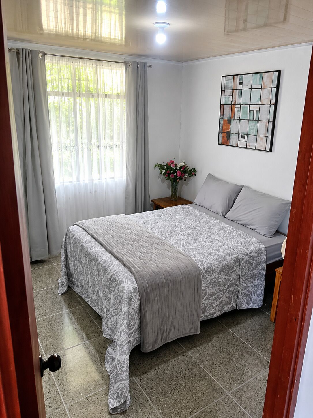 Une chambre à coucher avec un lit, des rideaux, une table de chevet ornée d’un vase de fleurs et une œuvre d’art accrochée au mur.