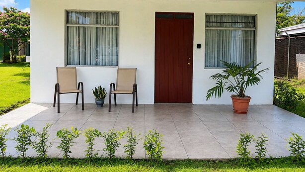 Une maison avec une porte rouge, deux fenêtres avec des rideaux, une plante en pot et un petit jardin.