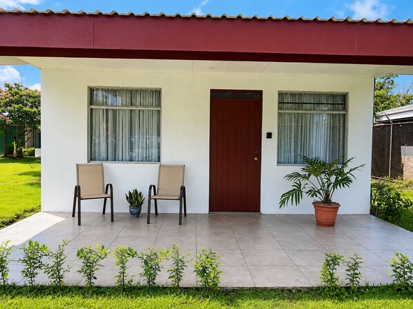 Une maison avec une porte rouge, deux fenêtres avec des rideaux, une plante en pot et un petit jardin.