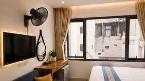 Habitació d'hotel amb llit, sofà, televisió, escriptori, ventilador i finestra amb vista a edificis.