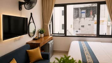 Habitación de hotel con cama, sofá, televisión, escritorio, ventilador y ventana con vista a edificios.