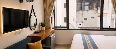 A hotel room with a bed, a sofa, a TV, a desk, a fan, and a window with a view of buildings.