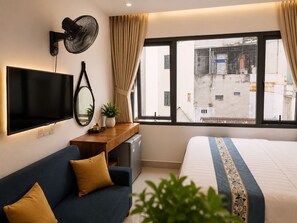 Ein Hotelzimmer mit Bett, Sofa, Fernseher, Schreibtisch, Ventilator und Fenster mit Blick auf Gebäude.