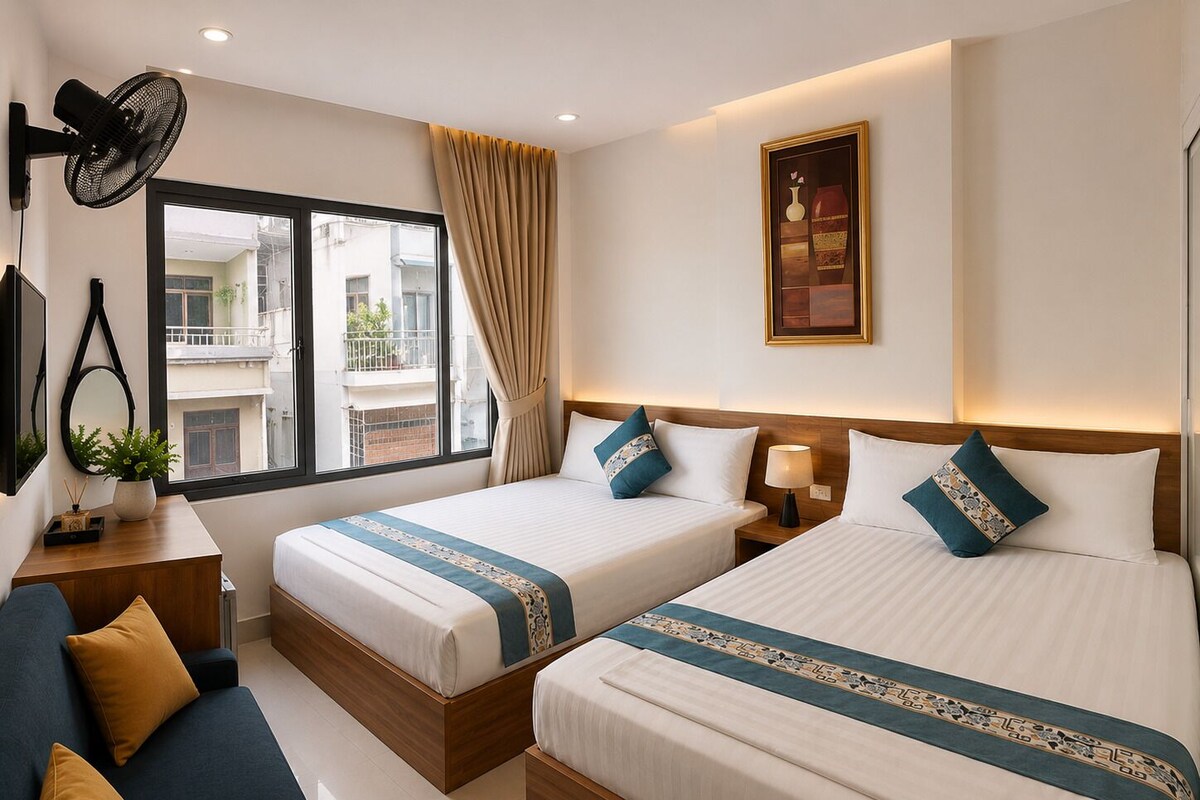 Ein Hotelzimmer mit zwei Betten, einem Sofa, einem Schreibtisch, einem Ventilator, einem Fenster mit Vorhängen und einem Wandbild.