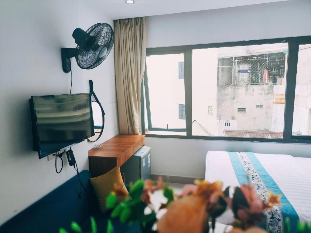 Et hotelværelse med en seng, et fjernsyn ophængt på væggen, et vindue med udsigt til bygninger og en ventilator på væggen.