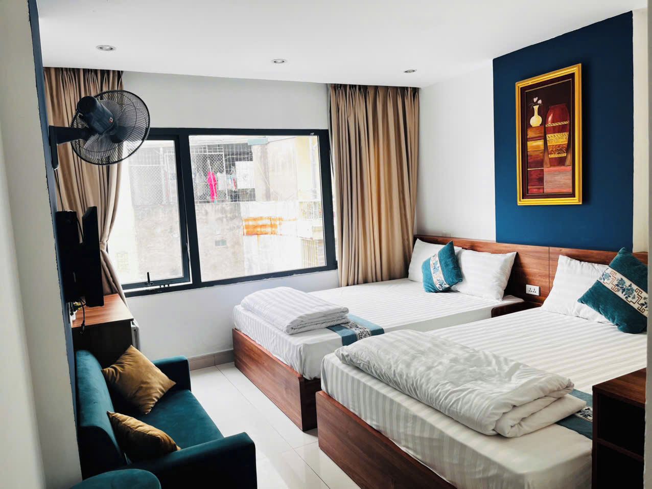 A hotel room with two beds, a sofa, a standing fan, and a large window with curtains.