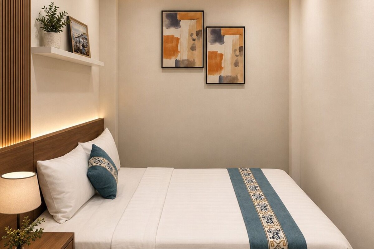 Ein Hotelzimmer mit einem Bett, Nachttisch, Lampe, einer an der Wand befestigten Ablage mit einer Pflanze und zwei gerahmten Kunstwerken.
