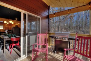 A wooden deck with a grill and chairs, a dining area with a table set for four, and a sliding glass door leading to a kitchen. - Private Indoor Pool, Hot Tub, Game Room, Sleeps 12 · 20 Min to Pigeon Forge (Sevierville)