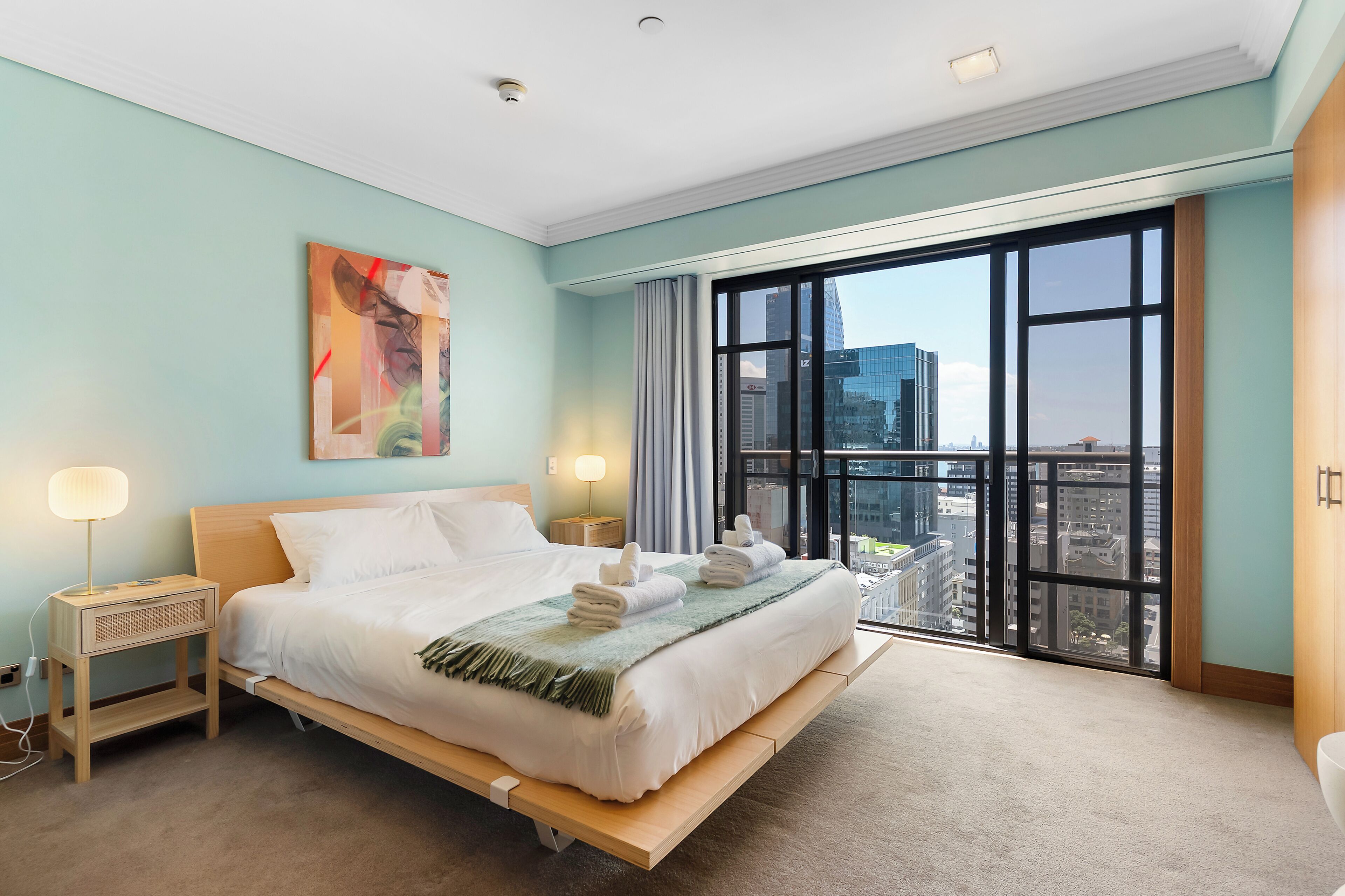 A hotel room with a large bed, bedside lamps, a painting, and a view of the city through large windows.