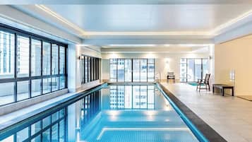 Piscina interior moderna com paredes de vidro transparente, piso ladrilhado e área de bancos.