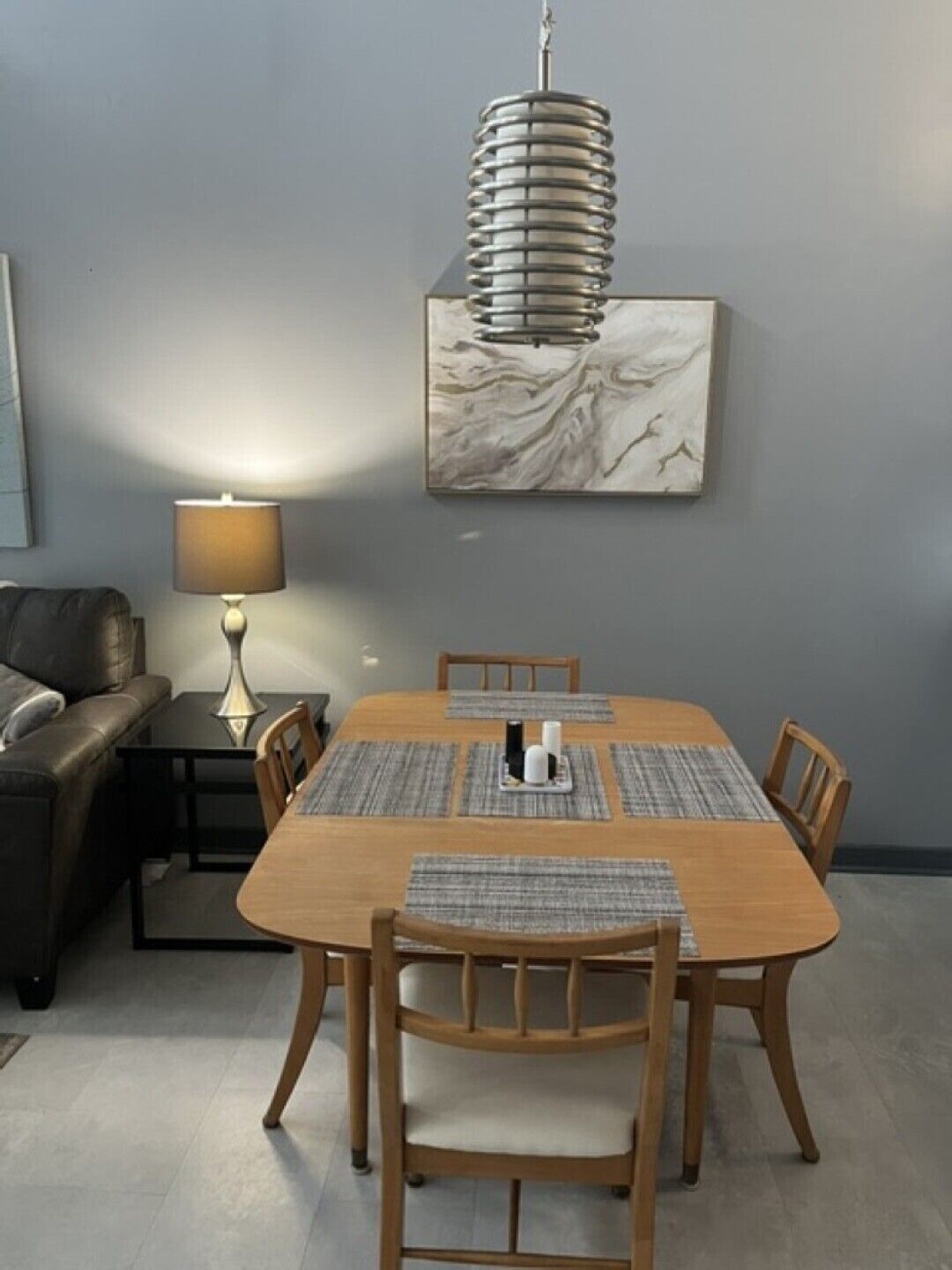Un comedor con una mesa de madera, seis sillas, una lámpara central, una obra de arte en una pared de mármol y una lámpara en una mesita auxiliar.