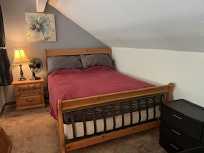 A bedroom with a wooden bed, a nightstand with a lamp, a fan, and a dresser.