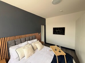 Een moderne slaapkamer met een bed, een houten hoofdbord, een klein tafeltje met glazen en een schilderij aan de muur.