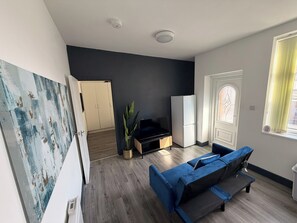 Een woonkamer met een blauwe bank, een televisie op een houten dressoir, een plant en een kledingkast.