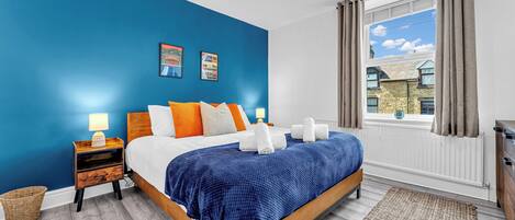 A bedroom with a blue wall, a wooden bed frame, white bedding, and a view of a building through a window with curtains.