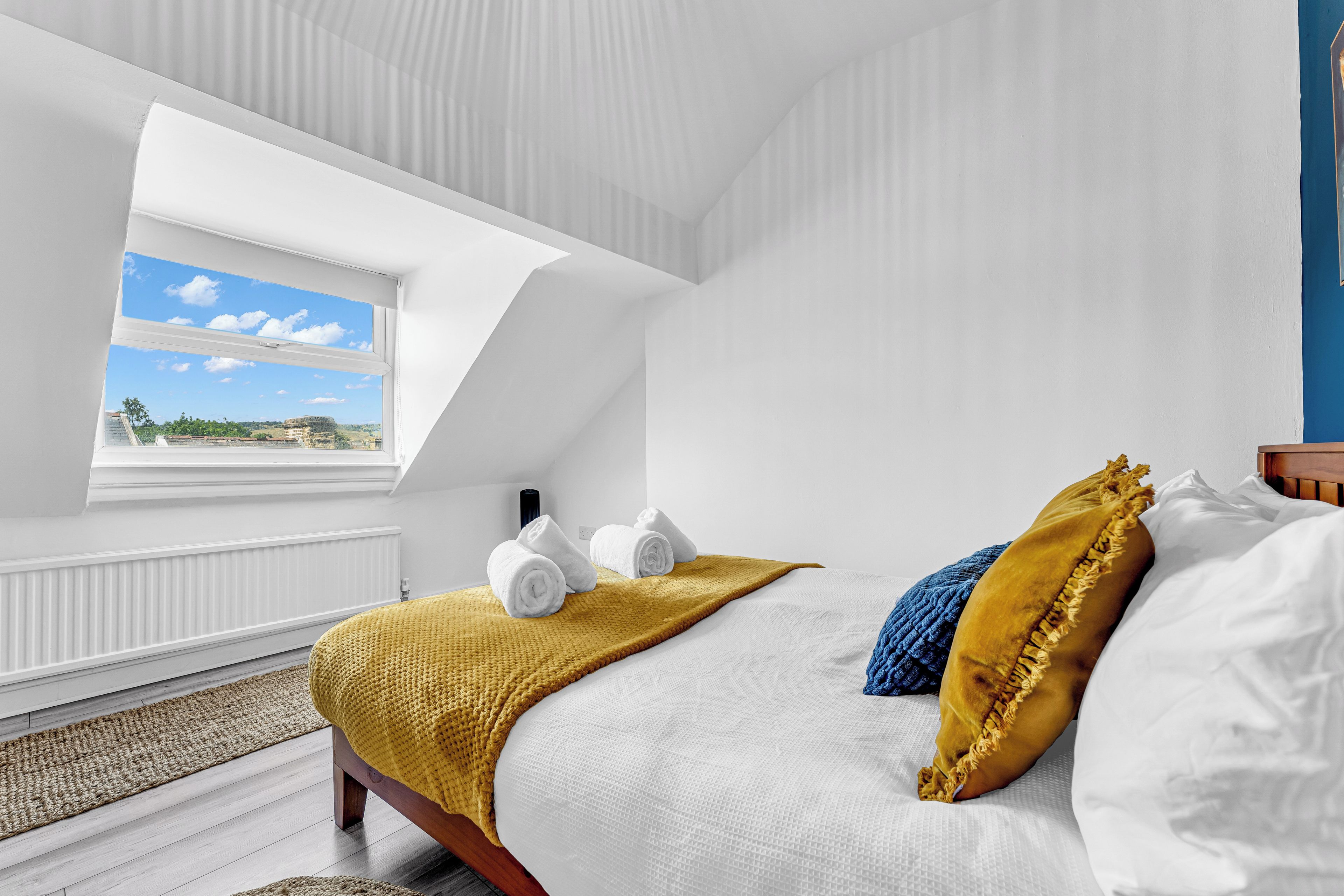 A bedroom with a skylight, a bed with white linens, a yellow throw blanket, and a view of the sky and buildings.