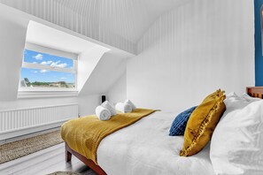 Ein Schlafzimmer mit Dachfenster, einem Bett mit weißen Leinen, einer gelben Decke und Blick auf Himmel und Gebäude.