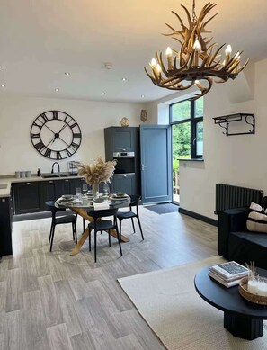 A modern kitchen with a large clock, a dining area with a round table and chairs, and a living area with a black sofa and a coffee table. - New Cosy Retreat in Matlock Bath (Matlock Bath)