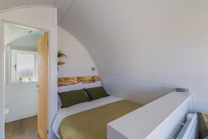 Quarto com teto inclinado, cabeceira de madeira, cama com roupa de cama verde e branca, uma pequena planta na parede e um radiador branco.