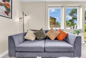 Ein graues Sofa mit vier Kissen in einem Raum mit einem großen Fenster, einer Stehlampe und einem gerahmten Bild an der Wand.