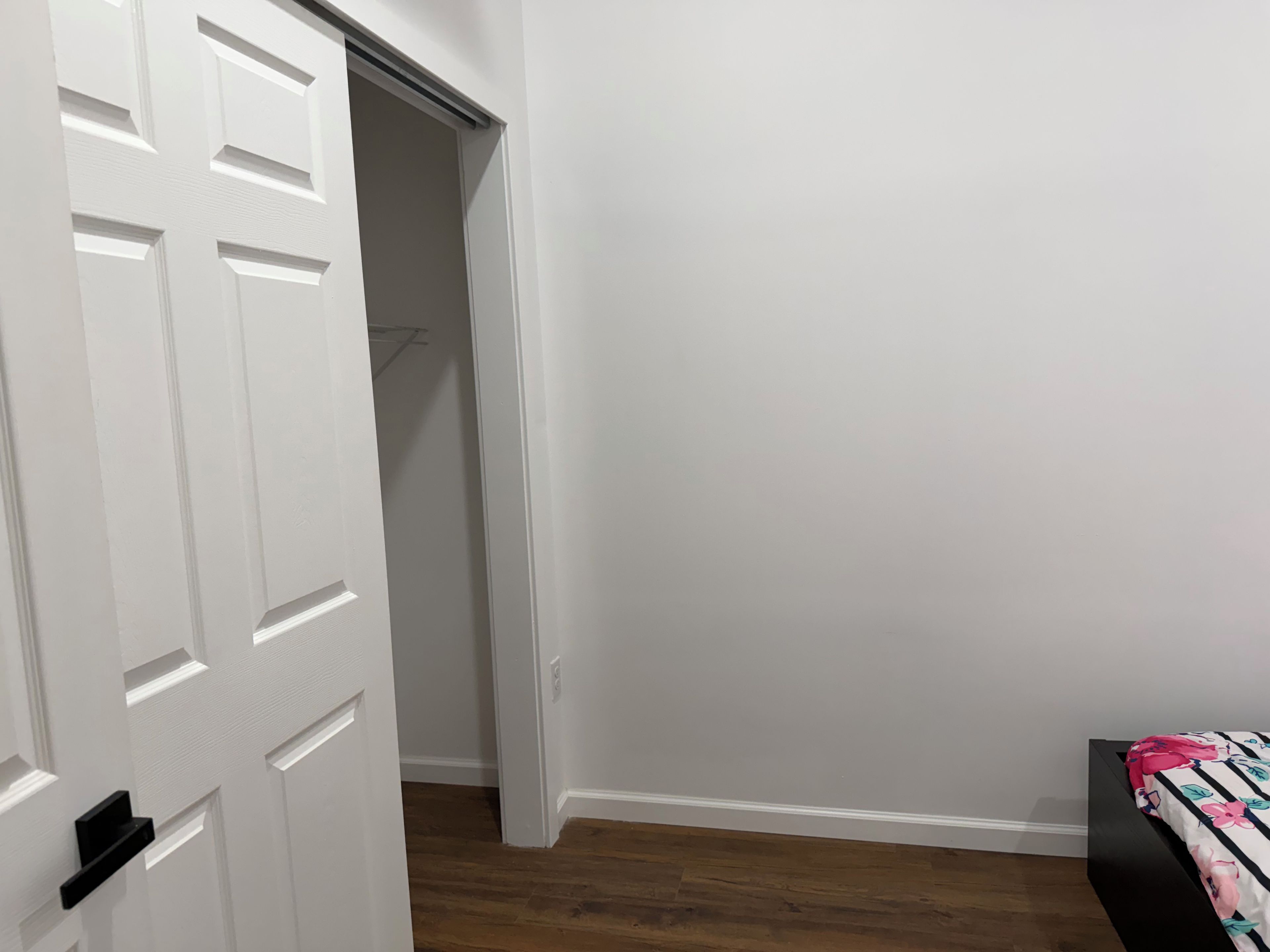 A room with a wooden floor, a bed with a floral bedspread, and a white door leading to a closet.