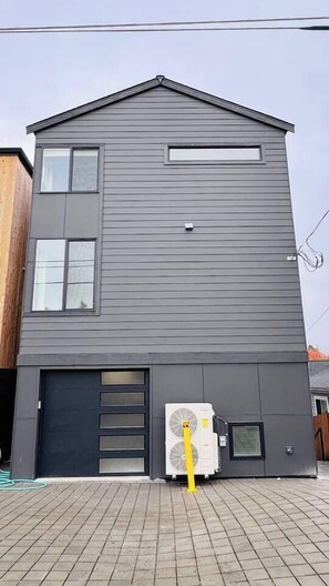 A two-story building with a dark grey exterior, a garage door, and a large air conditioning unit in front. - Premium 3BR Suite Minutes to Downtown (Seattle)