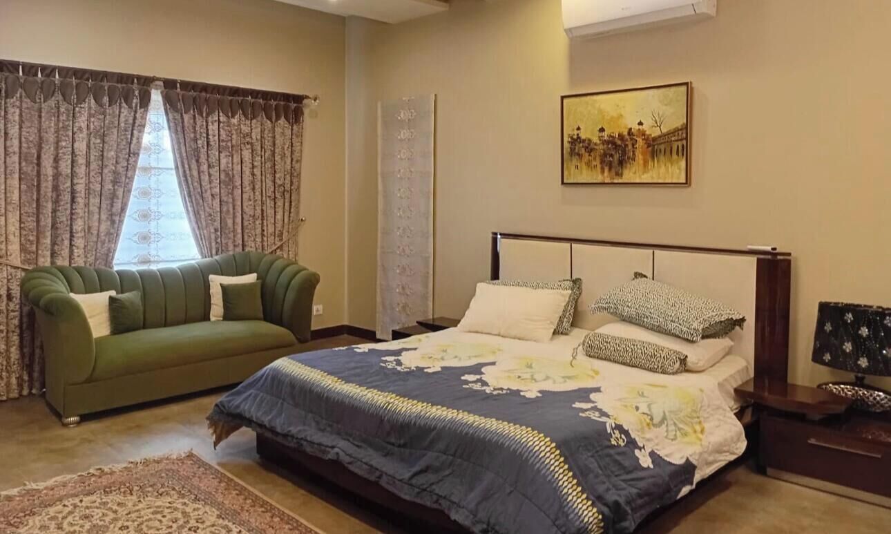 Un dormitorio con cama, sofá verde, alfombra estampada, un cuadro en la pared y ventana con cortinas.