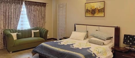 A bedroom with a bed, a green sofa, a patterned rug, a painting on the wall, and a window with curtains.